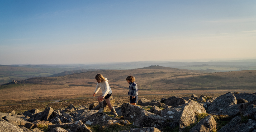 Two people hike across rocky moorland on a sunlit hillside, with rolling fields and a hazy horizon stretching out behind them.